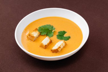 Kahverengi arka planda beyaz bir tabakta kızarmış ekmek ve otlu portakal kremalı balkabağı çorbası.