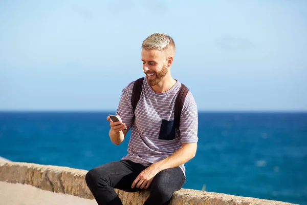 携帯電話を見て海辺に座って幸せな若い男の肖像 — ストック写真