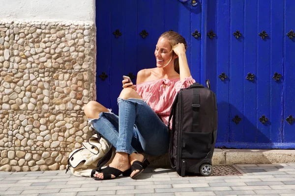 Portrait of travel woman sitting on sidewalk with suitcase and mobile phone