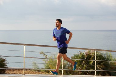 Deniz kıyısında koşan sağlıklı genç adam tam uzunlukta yan portresi