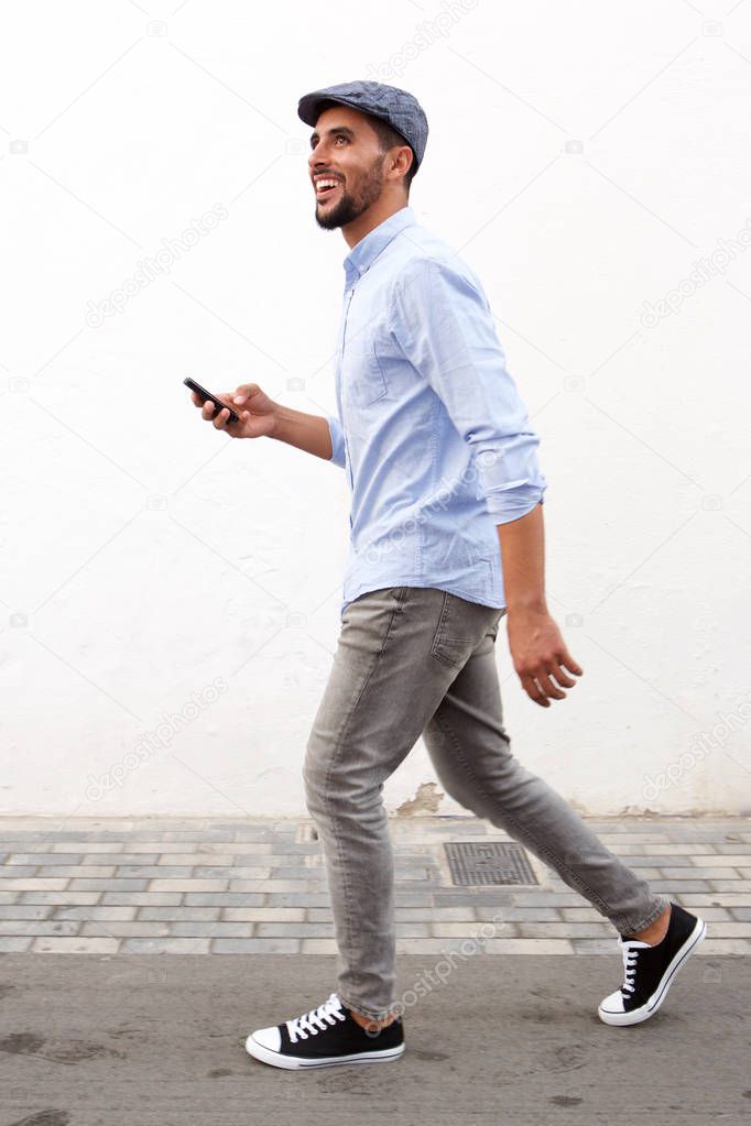 Full length side portrait of smiling man walking with cellphone against white wall