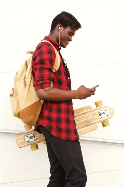 スケート ボード 携帯電話を見てバック パックとハンサムな若いアフリカ男の側ビュー肖像画 — ストック写真