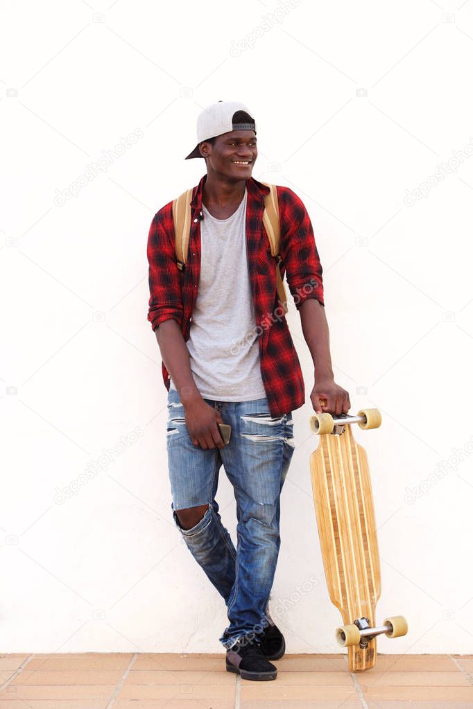 Full length portrait of smiling young guy standing against wall with skateboard