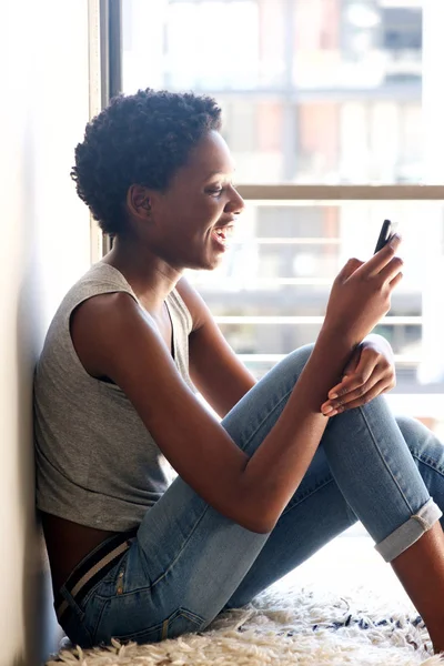 Side portrait of smiling young black woman using smart phone by window at home