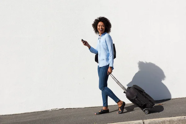 Full body portrait of laughing young female traveler walking with suitcase and mobile phone