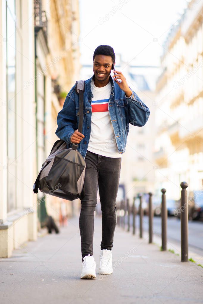 Full length portrait of happy young african american man walking with bag and talking with cell phone in city