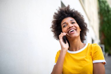 Beyaz duvarda cep telefonuyla konuşan mutlu Afro-Amerikan genç kadın portresi.