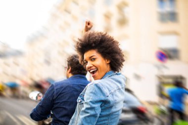 Scooter 'ın arkasında oturan mutlu Afro-Amerikan kadının portresi şehir sokaklarında kolları havada geziyor.