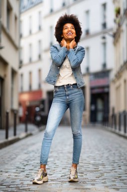 Gülümseyen Afro-Amerikan kadın modelin tüm vücut portresi şehirde duruyor.