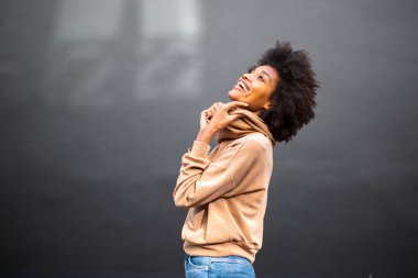 Gri duvarda kahkahalar atan afro saçlı çekici siyah bir kadının yan portresi.
