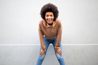 Afro saçlı genç siyahi bir kadının portresi. Gri duvarın yanında elleri dizlerinde gülüyor.