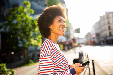 Şehirde cep telefonuyla duran mutlu Afro-Amerikan kadının yan portresi.