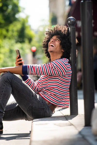 Portret Szczęśliwego Młodzieńca Siedzącego Ulicy Telefonem Komórkowym Śmiejącego Się Patrząc — Zdjęcie stockowe