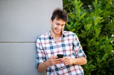 Duvara yaslanmış cep telefonuna bakan gülümseyen genç adamın portresi 