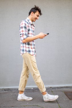 Kaldırımda yürüyen ve cep telefonuna bakan gülümseyen genç adamın tam boy yan portresi.