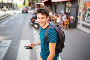 Gülümseyen genç adam çantasıyla ve cep telefonuyla şehir caddesinde yürüyor.
