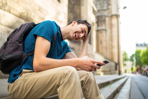 携帯電話やバッグで街の外に座って幸せな若い男のサイドポートレート — ストック写真