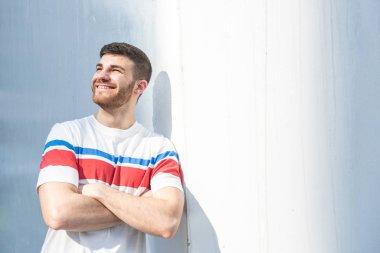 Kollarını kavuşturup beyaz duvara yaslanan gülümseyen genç adamın portresi.