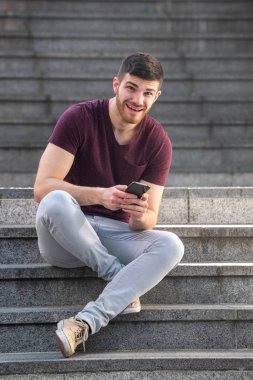 Dışarıda merdivenlerde cep telefonuyla oturan genç adamın portresi.