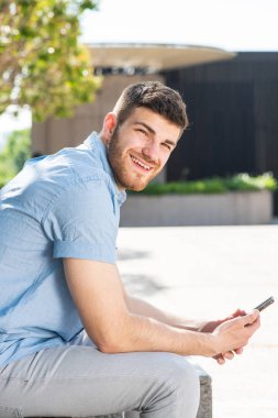 Dışarıda cep telefonuyla oturan mutlu genç adamın yan portresi.