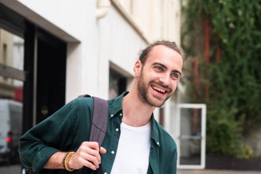 Sakallı ve sırt çantalı bir gencin portresi. Dışarıda gülüyor.