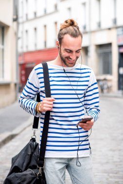 Portre. Gülümseyen genç adam şehirde yürüyor. Akıllı telefon ve kulaklıkla müzik dinliyor.