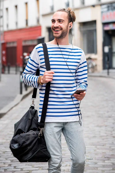 スマートフォンやイヤホンで音楽を聴きながら街を歩くポートレート笑顔の旅行男 — ストック写真