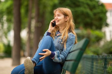 Park bankında oturmuş cep telefonuyla konuşan gülümseyen genç bir kadının yan portresi.