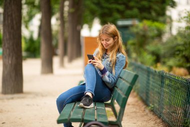 Park bankında oturan ve cep telefonuna bakan genç bir kadının portresi.