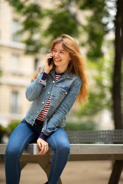 Parkta oturmuş cep telefonuyla konuşan mutlu genç bir kadının portresi.