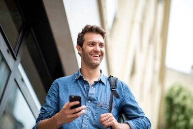 Şehirde cep telefonu ve çantasıyla gülümseyen bir adam var.