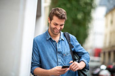 Duvara yaslanmış cep telefonuna bakan gülümseyen adamın portresi.