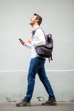 Tam vücut portresi gülümseyen adam cep telefonu ve çantasıyla gri duvarda yürüyor.