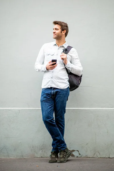 フルボディポートレートのハンサムな男とともに携帯電話とバッグによってグレーの壁 — ストック写真