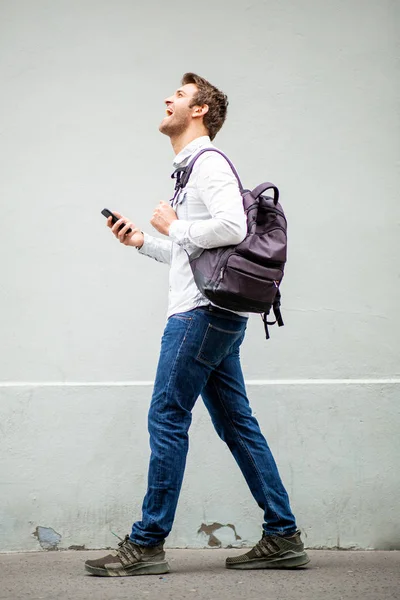 フルボディ側の肖像画笑顔男歩くとともに携帯電話とバッグによってグレーの壁 — ストック写真
