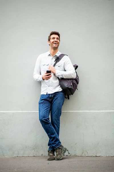 フルボディポートレートの幸せなハンサムな男とともに携帯電話とバッグによってグレー壁 — ストック写真