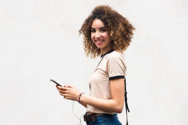 Cep telefonu ve kulaklıkları beyaz arka planda olan gülümseyen genç bir kadının yan portresi