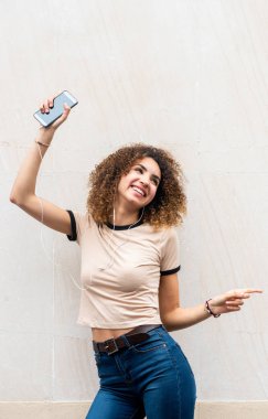 Duvarda müzik dinlerken cep telefonu ve kulaklıklarla dans eden neşeli Afro-Amerikan kadın portresi.