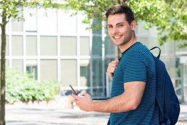 Kentin dışında çantalı ve cep telefonlu gülümseyen genç adamın yan portresi.