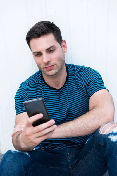一个英俊的年轻人坐在白墙边看手机的画像 — 图库照片