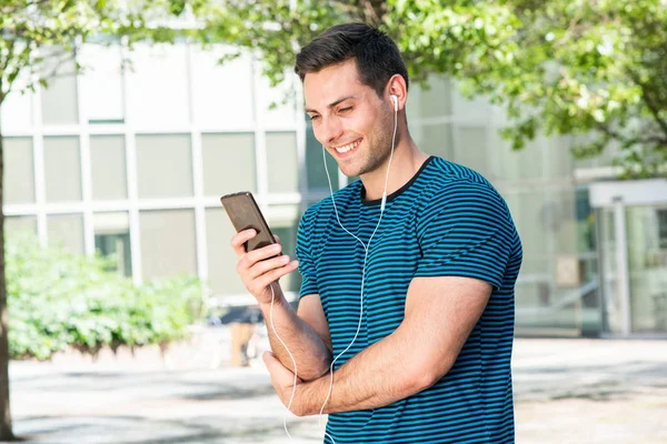 Portrét Usmívající Mladý Muž Při Pohledu Mobilní Telefon Sluchátky Venku — Stock fotografie