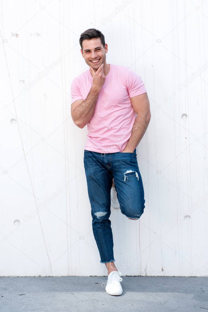 Full length portrait of cool young man leaning against white wall and smiling