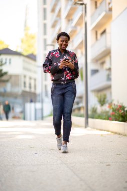 Full body portrait smiling young african woman walking in city looking at mobile phone