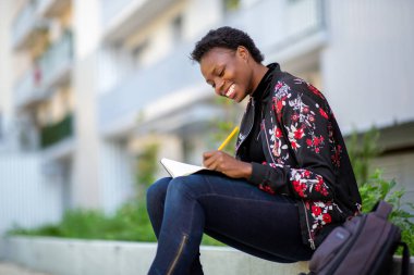 Side portrait smiling young african woman sitting outside with book 