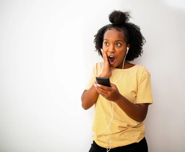 Portrait surprised african American girl with mobile phone and earphones against white background