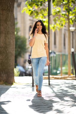 Full length portrait happy young woman walking and talking with cellphone in city