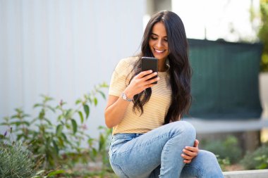 portrait of smiling young woman sitting outside looking at cellphone