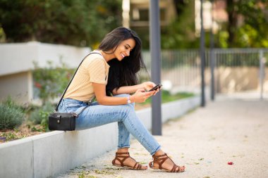 Full length side portrait young woman sitting outside smiling looking at mobile phone