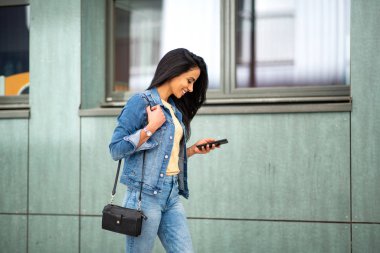 Side portrait of smiling young hispanic woman walking with mobile phone and purse