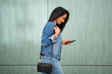 Side portrait of smiling young latin woman walking with mobile phone and purse